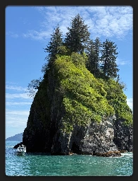 land formation jetting our of the ocean. 
looks like a very small but extremely tall island. teal ocean water touching black rocks that make up the base. green vegetation stretches up & pine trees top the island like candles on a cake.  A boat sits in the water on the left. 